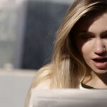 A young woman with blonde hair expresses surprise while reading a newspaper outdoors in daylight.