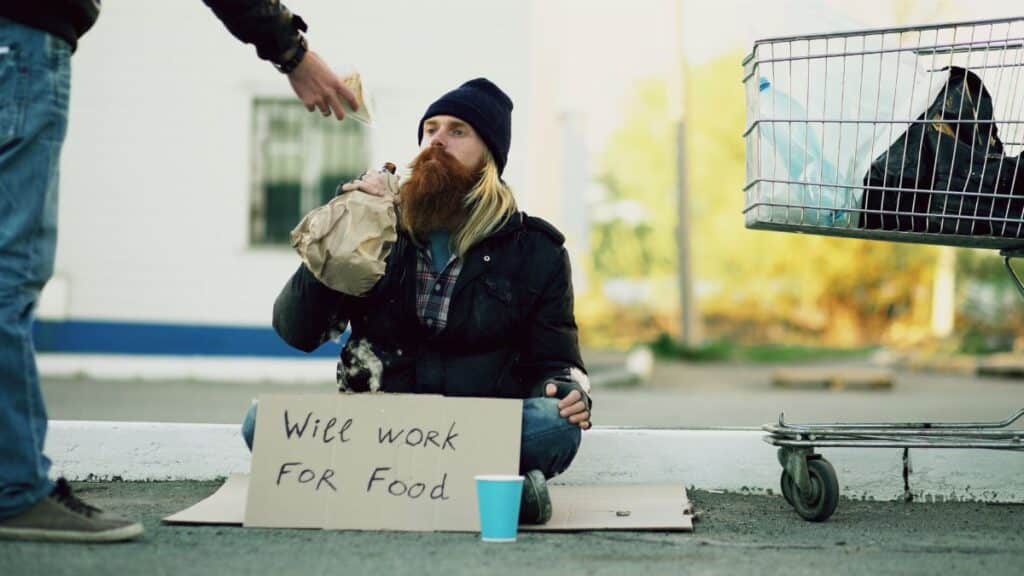 Homeless individual with 'Will Work For Food' sign receives help outdoors.