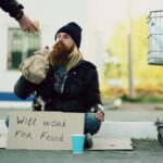 Homeless individual with 'Will Work For Food' sign receives help outdoors.