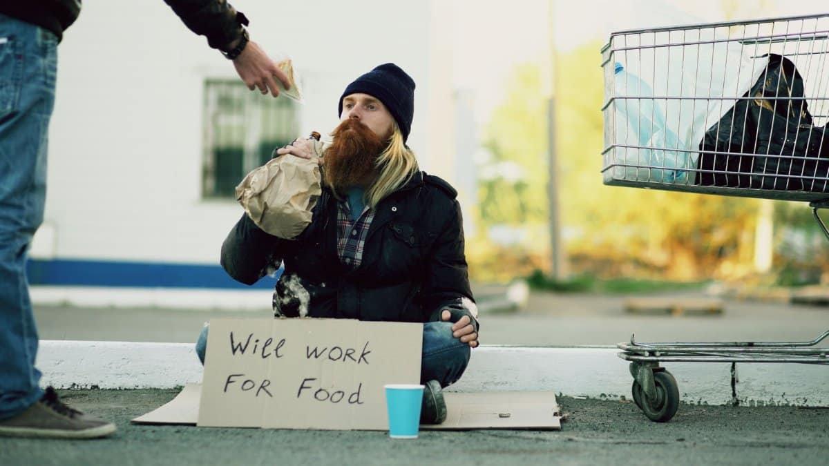 Homeless individual with 'Will Work For Food' sign receives help outdoors.