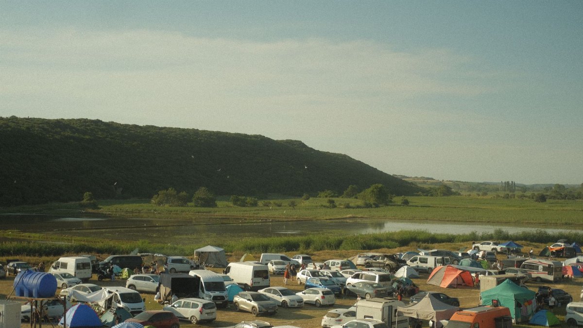 A lively camping festival with cars and tents by a scenic lake and mountains.