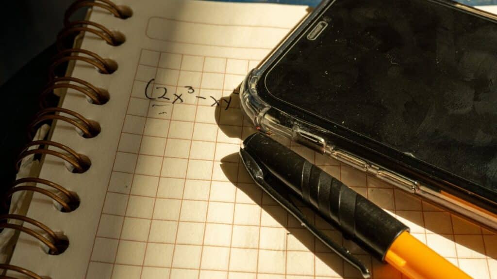 A notebook with a math equation, smartphone, and pen on checkered paper in warm lighting.