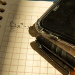A notebook with a math equation, smartphone, and pen on checkered paper in warm lighting.