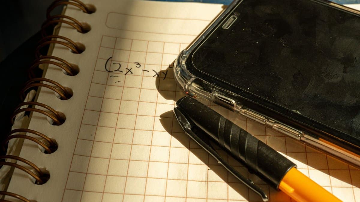 A notebook with a math equation, smartphone, and pen on checkered paper in warm lighting.
