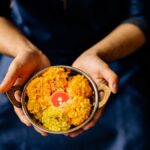 Hands holding a brass bowl with marigold flowers and a candle, symbolizing a Diwali ceremony.