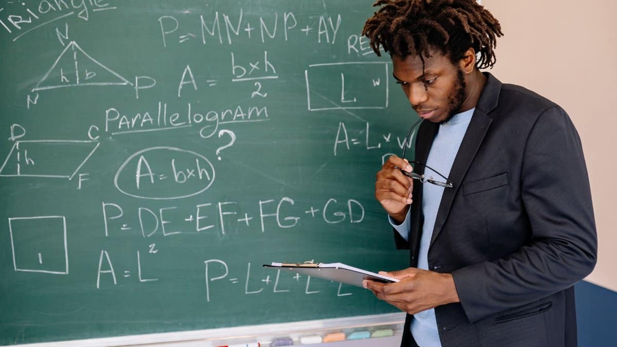 Educator in suit jacket solving math equations on blackboard, holding clipboard.