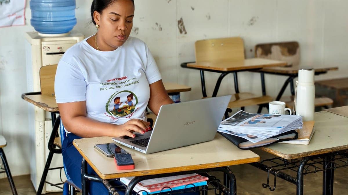 A woman in a classroom typing on a laptop, surrounded by educational materials, conveying a focus on work and study.