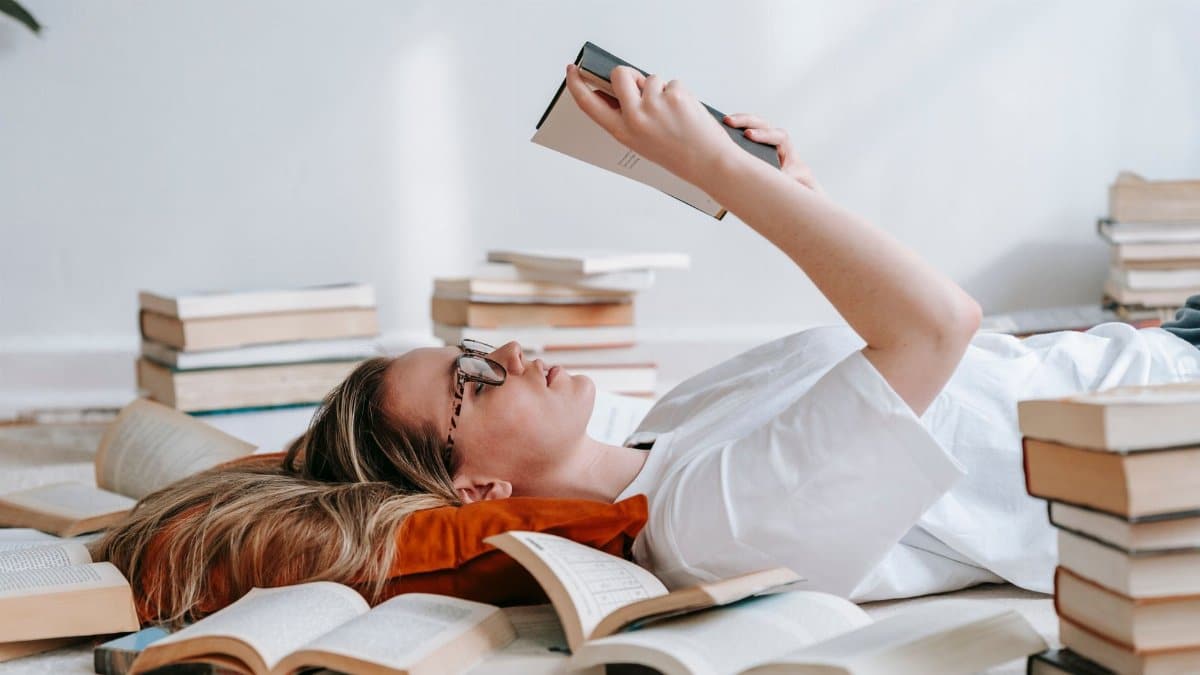 Side view of concentrated female lying among stacks of different literature and reading book for research