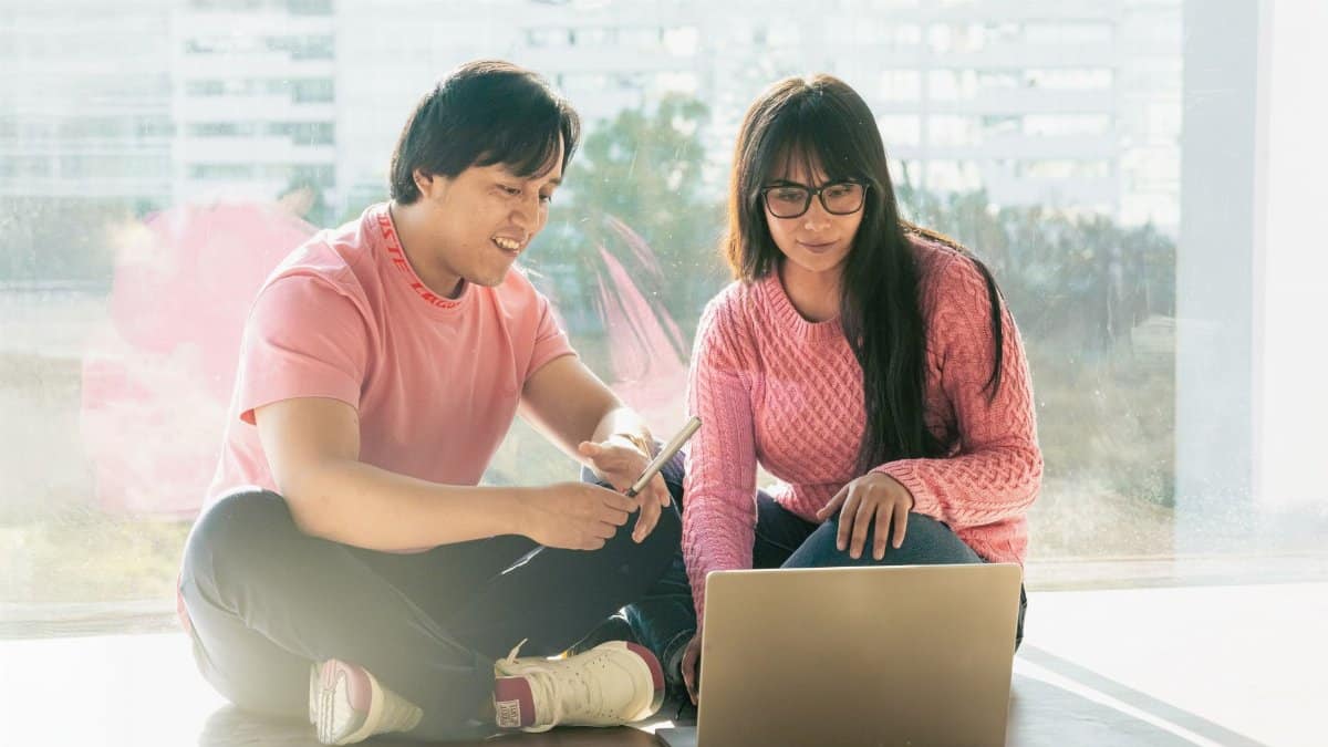 Two young adults using a laptop by a large window, engaging in tech-related activities.