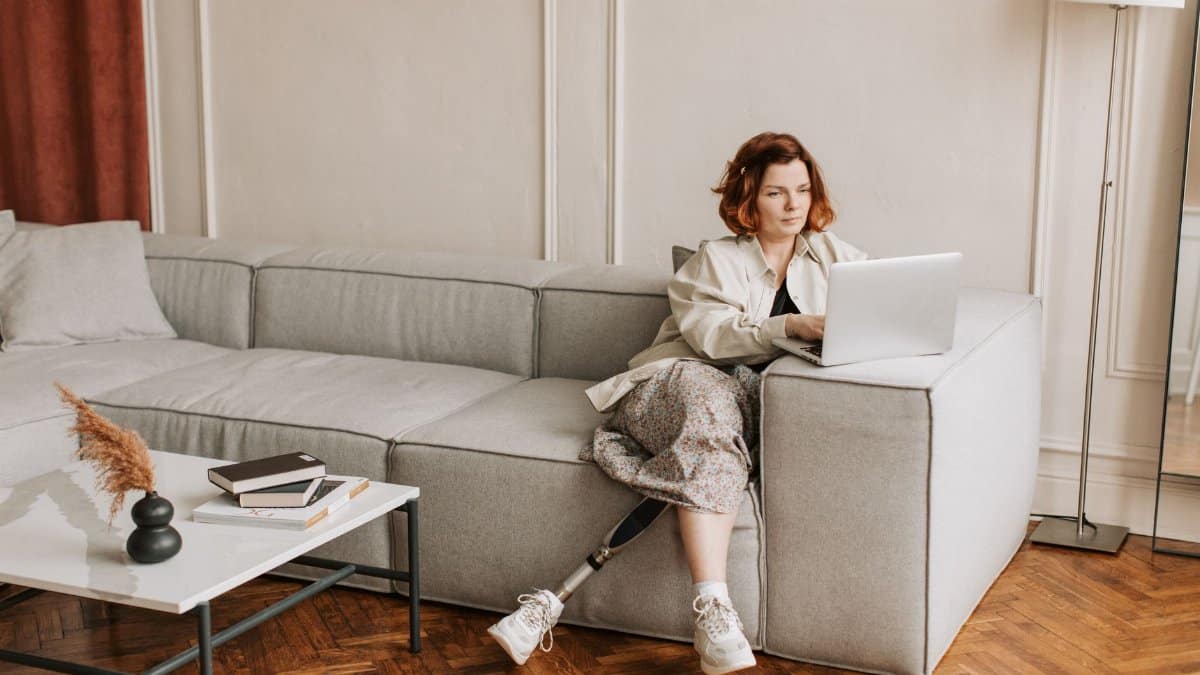 A woman with a prosthetic leg works remotely on a laptop in a cozy living room setting.