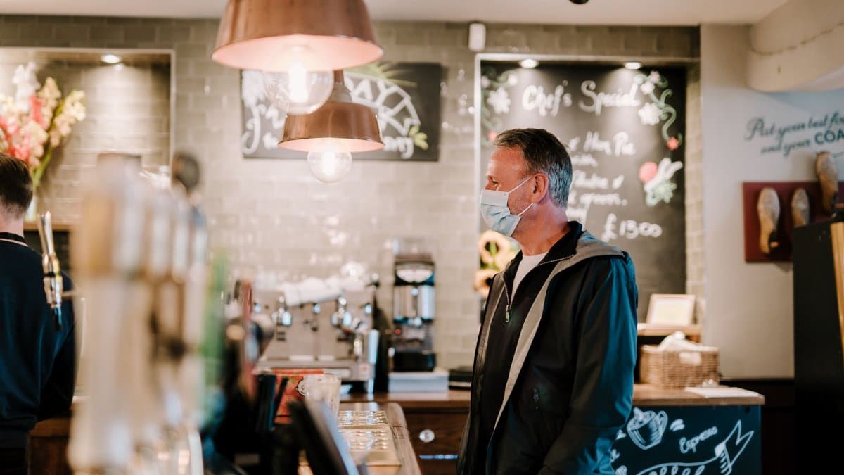 A masked man stands at a café counter, embracing COVID-19 precautions.