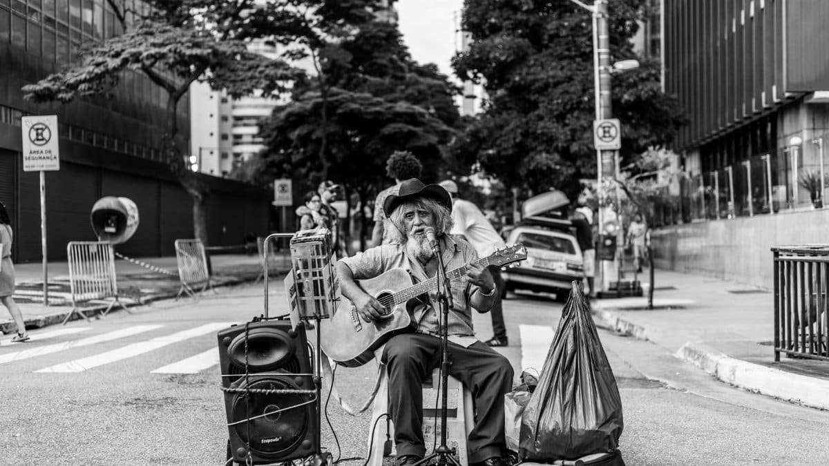 Black and white photo of an elderly street musician playing an acoustic guitar on a city road.