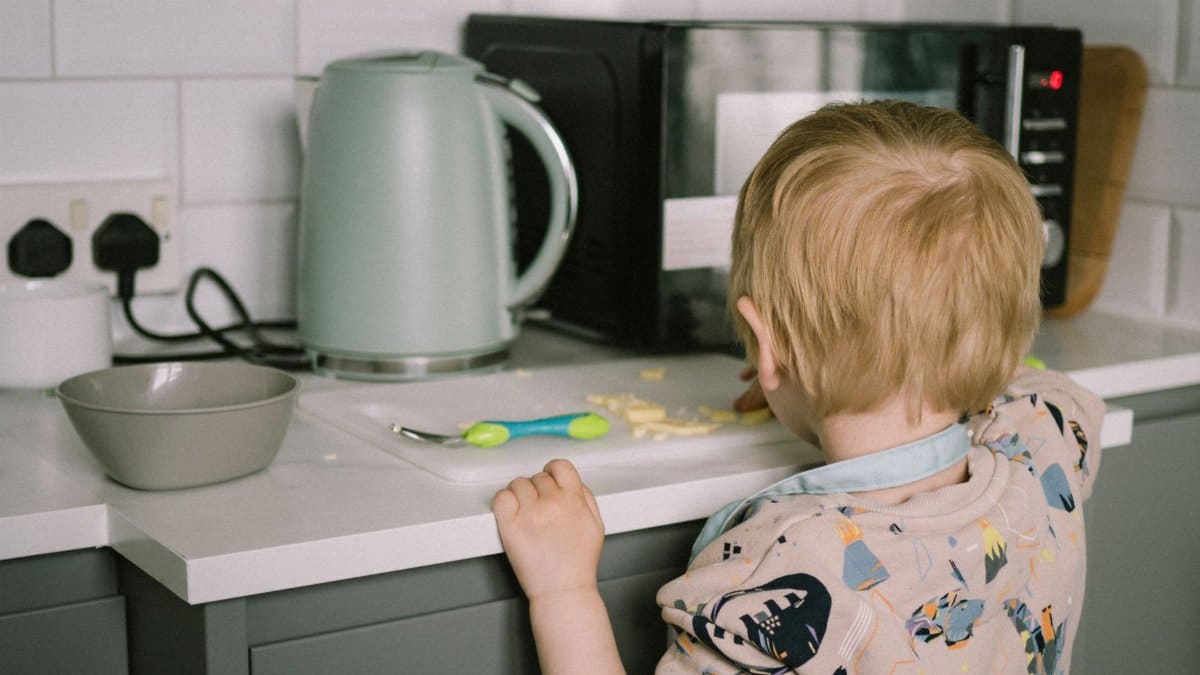 A toddler reaching for food on the kitchen counter next to a kettle and microwave.