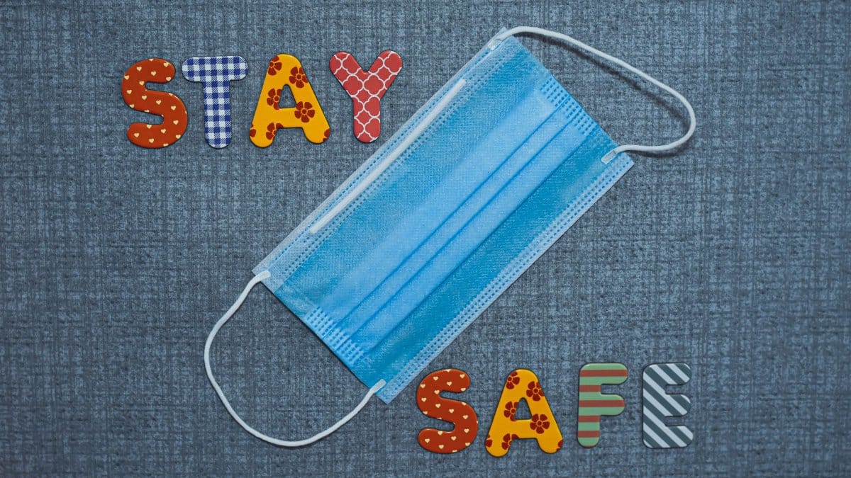 Colorful letters spelling 'Stay Safe' with a blue face mask on fabric background.