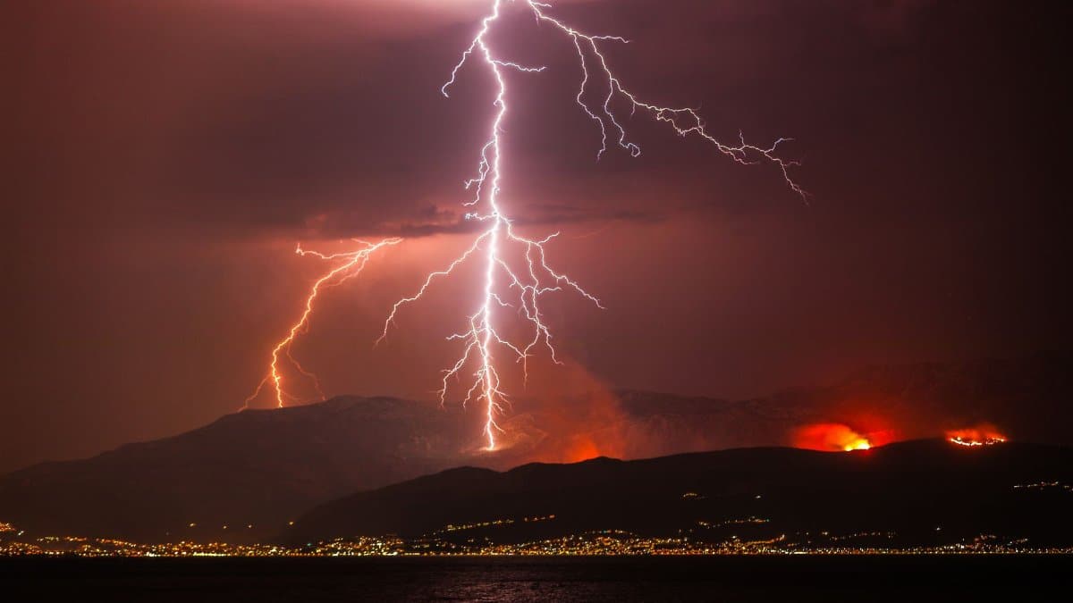 Lightning illuminates the night sky over a mountainous landscape with distant wildfires and city lights.