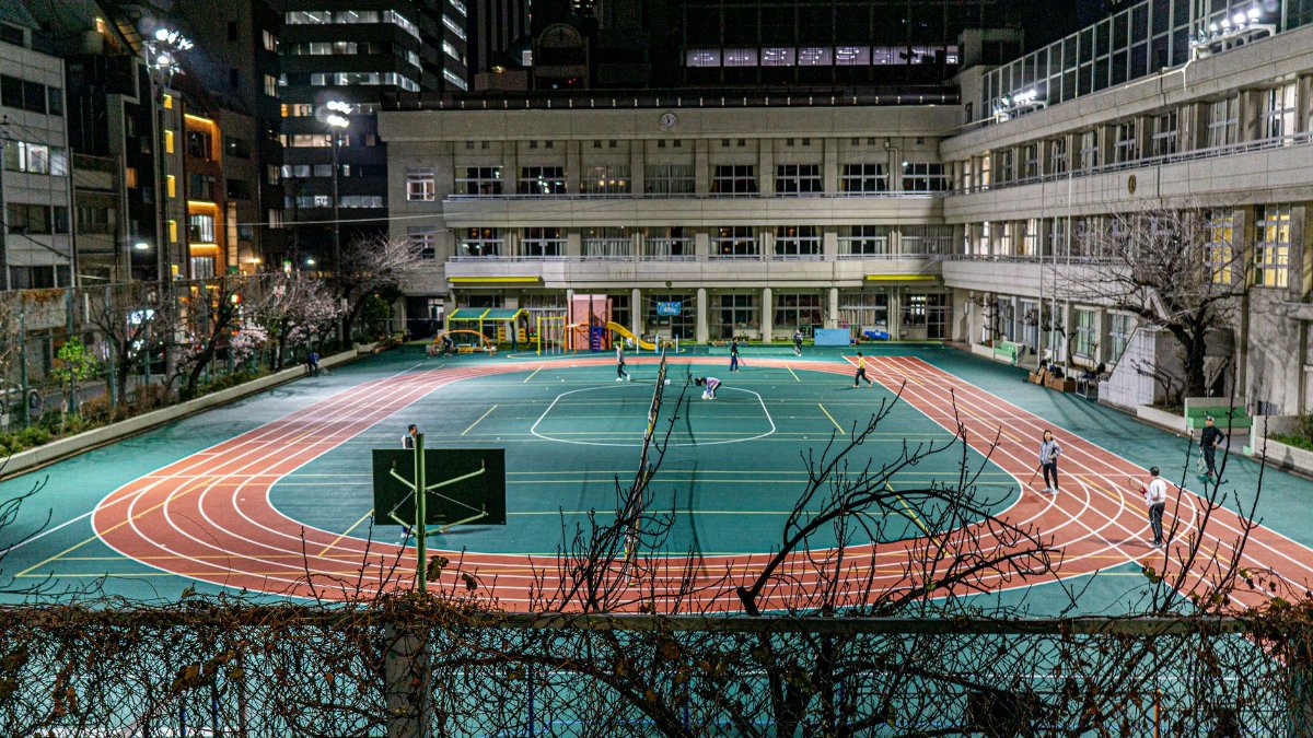 Nighttime view of urban school playground and track surrounded by buildings, with children playing.