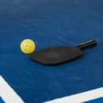A pickleball paddle and yellow ball on a blue court corner, symbolizing sport and activity.