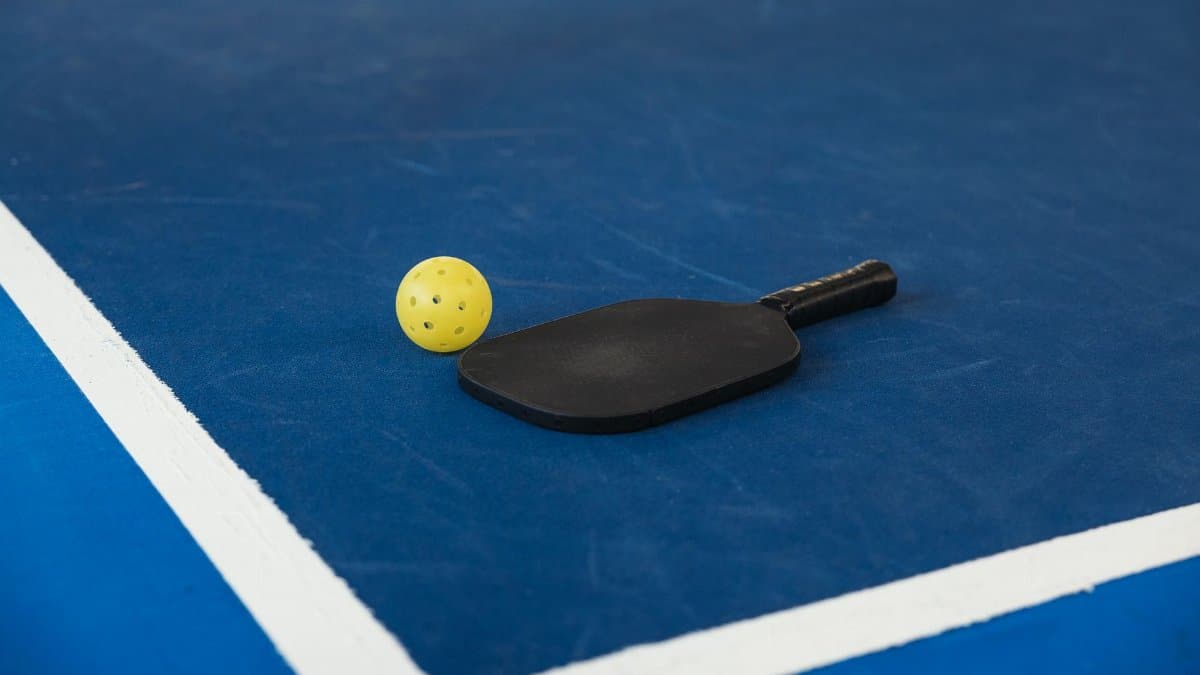 A pickleball paddle and yellow ball on a blue court corner, symbolizing sport and activity.
