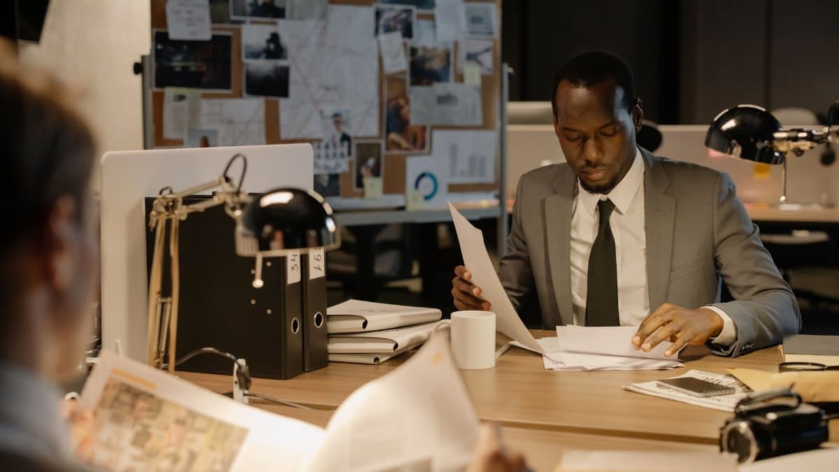 Detective in a suit examines papers at desk in a dimly-lit office, suggesting investigation work.