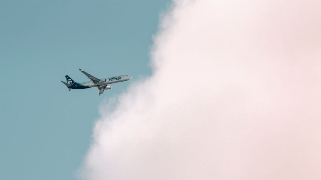 Alaska Airlines plane soaring through a bright sky with fluffy clouds in Seattle.