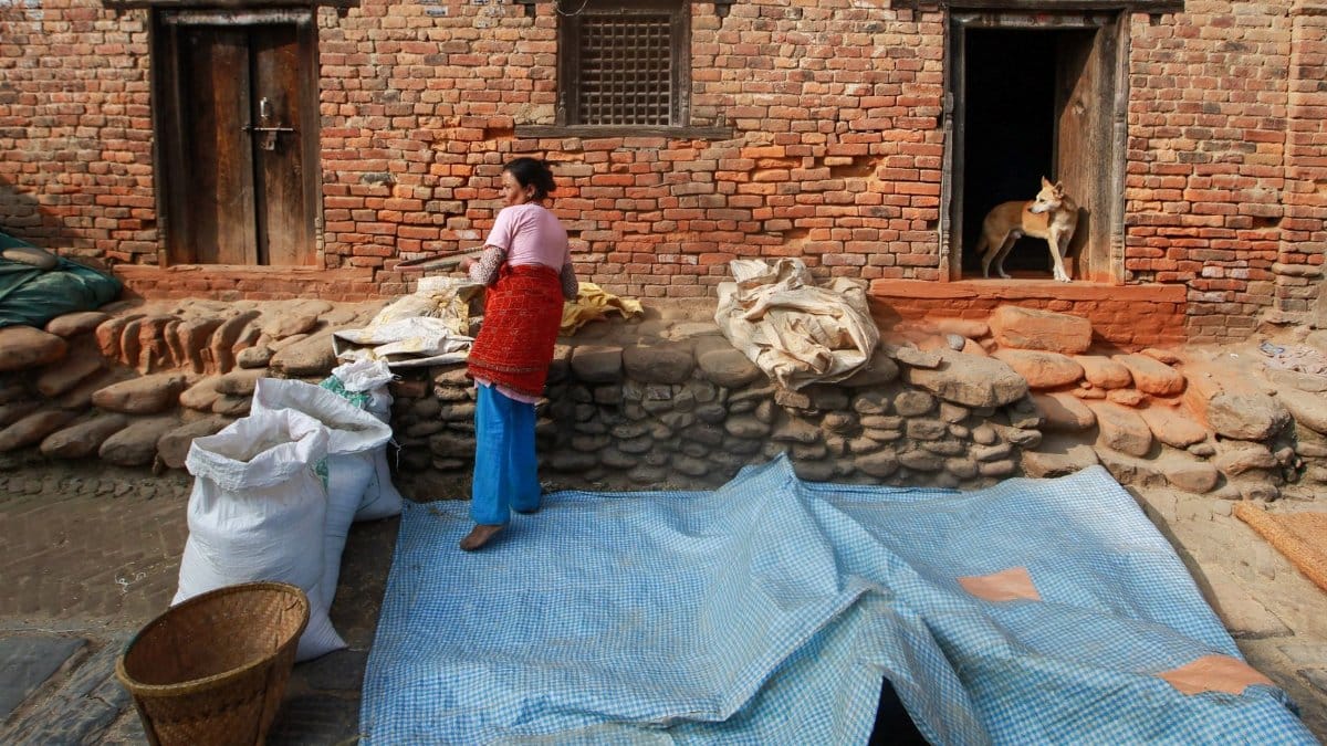 Woman handling grains outside a rustic brick house with storage bags and a dog nearby.