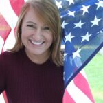 A woman smiling warmly while holding an American flag outside on a sunny day.