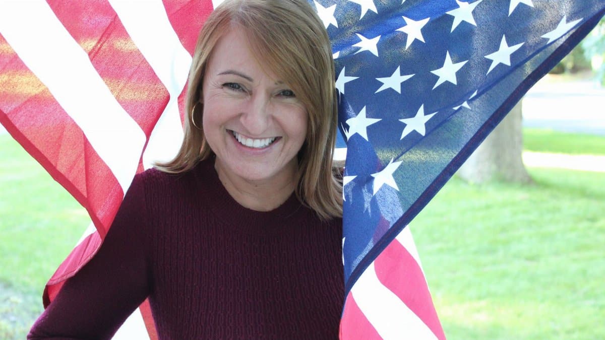 A woman smiling warmly while holding an American flag outside on a sunny day.