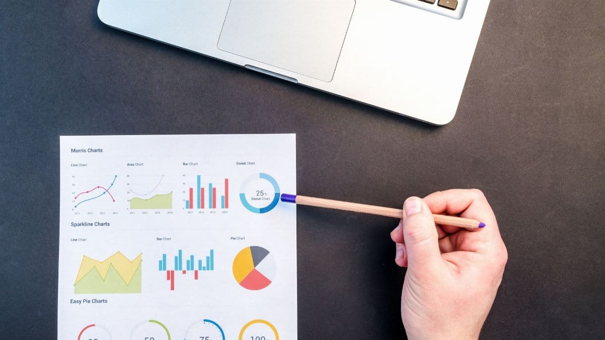 Hand holding pencil reviewing colorful data charts on desk with laptop.