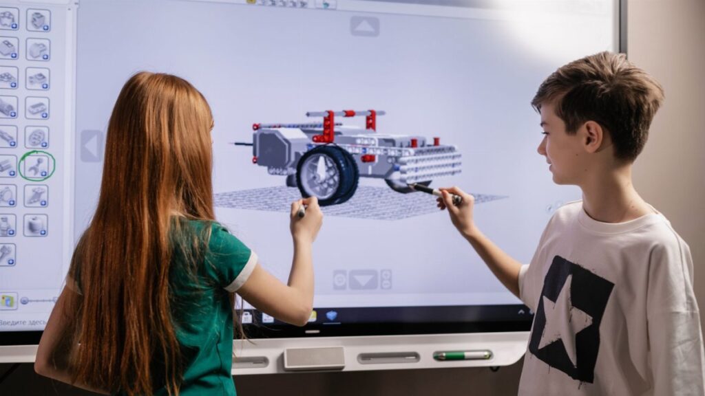 A boy and girl collaborate on a robotics project using a digital screen in a classroom setting.