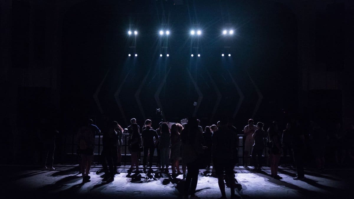 Silhouettes of a crowd gathered in front of a stage illuminated by bright lights at a live music event.
