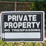 Close-up of a private property no trespassing sign on a chain-link fence with greenery in the background.