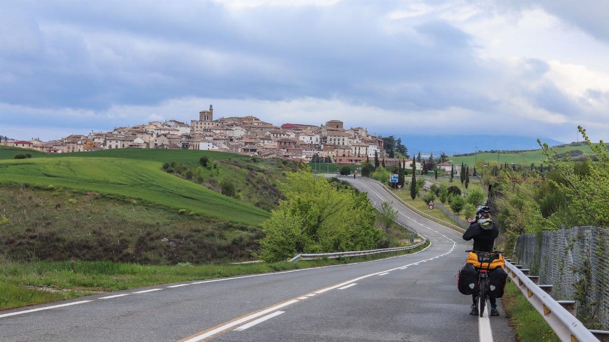 Breathtaking view of a cyclist on a winding road approaching a Spanish hilltop town under a moody sky.