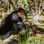 A man kneeling and attending to a grave at a sunlit cemetery.