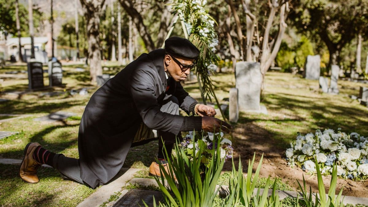 A man kneeling and attending to a grave at a sunlit cemetery.