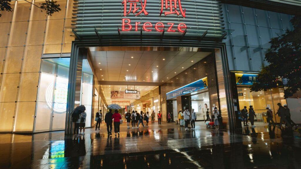 A bustling shopping mall entrance with people and umbrellas on a rainy evening.