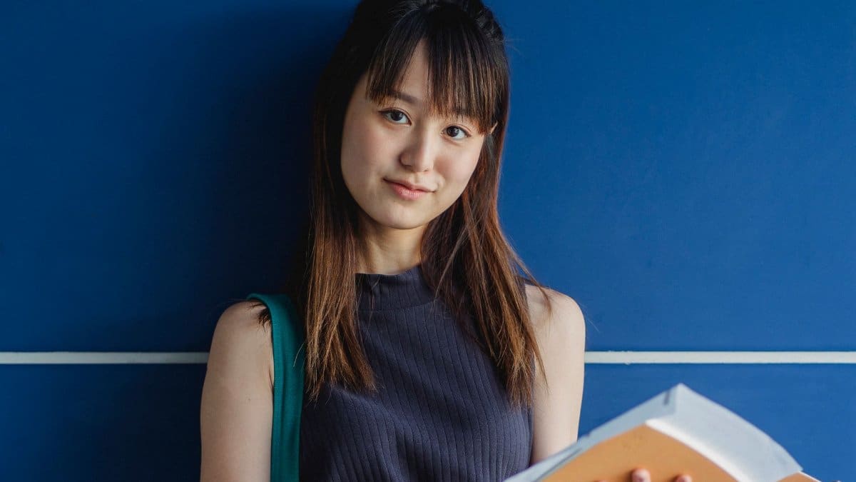 Young smiling ethnic woman with tilted head and workbooks looking at camera near tiled wall
