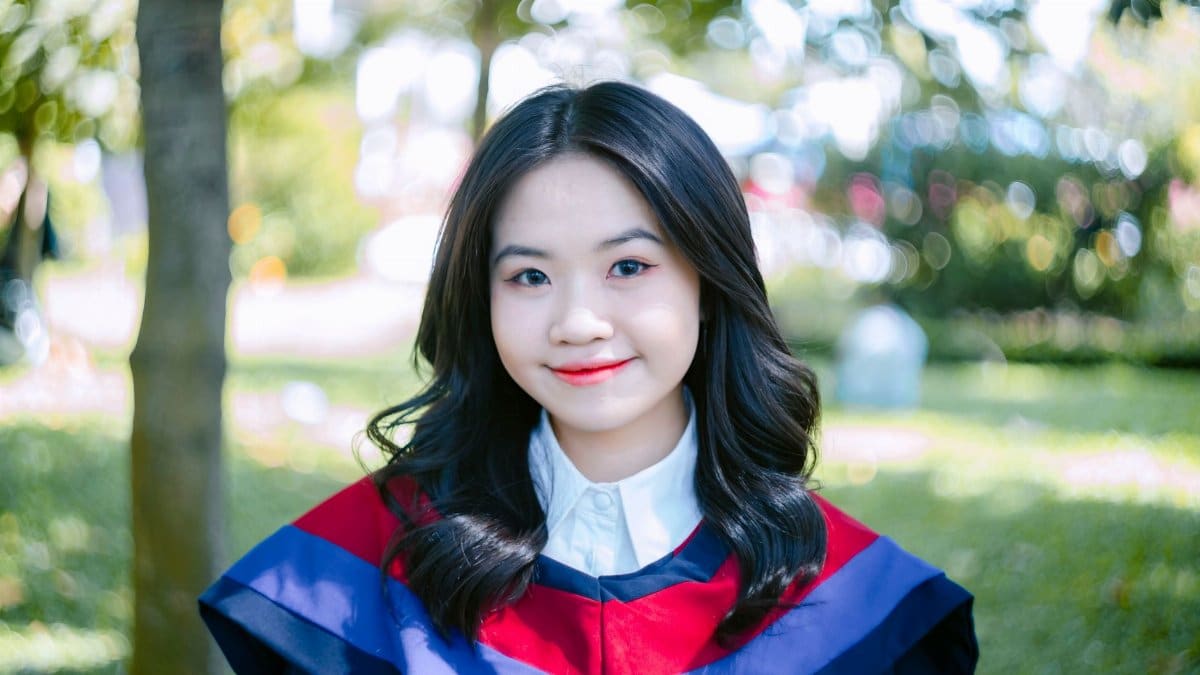 Portrait of a young Asian woman smiling in a graduation gown with a blurred park background.
