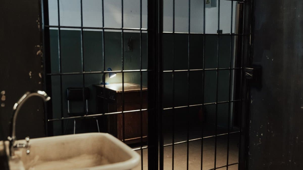 A dimly lit prison cell interior with metal bars, a sink, and a desk.