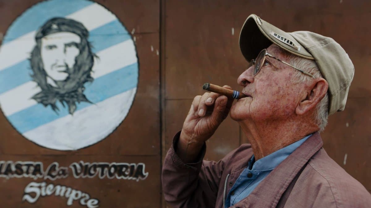 Senior man smoking a cigar in front of a mural featuring Che Guevara.