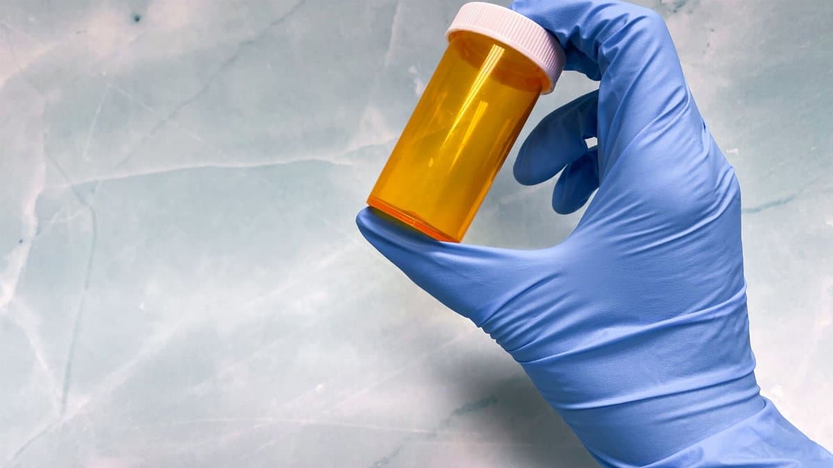 Close-up of a gloved hand holding an empty yellow prescription vial against a gray background.
