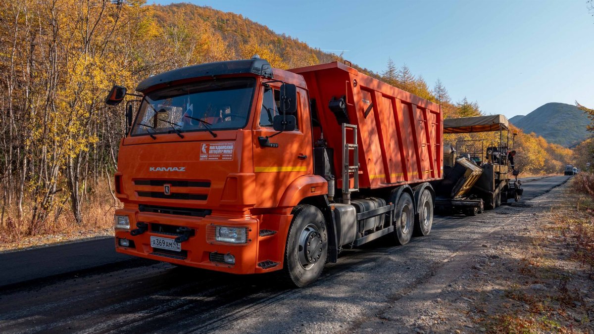 Orange truck and paver working on road construction in an autumn forest with vibrant fall foliage.