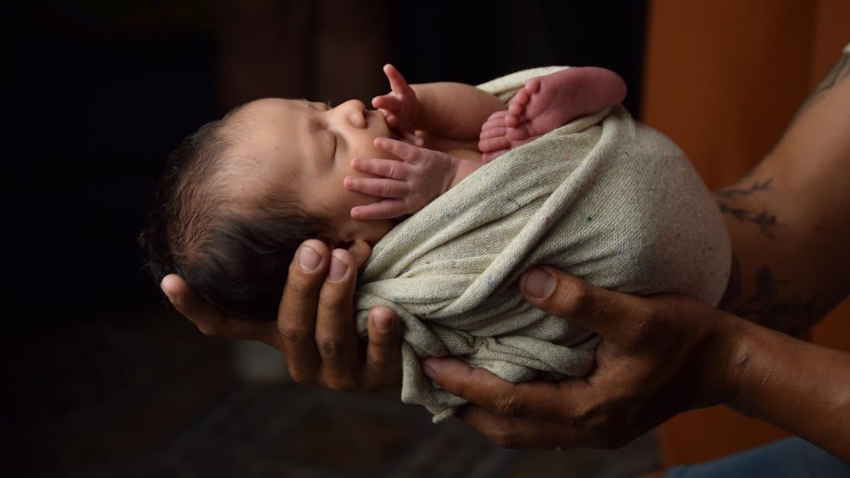 Newborn baby gently cradled in adult hands, swaddled in soft fabric.