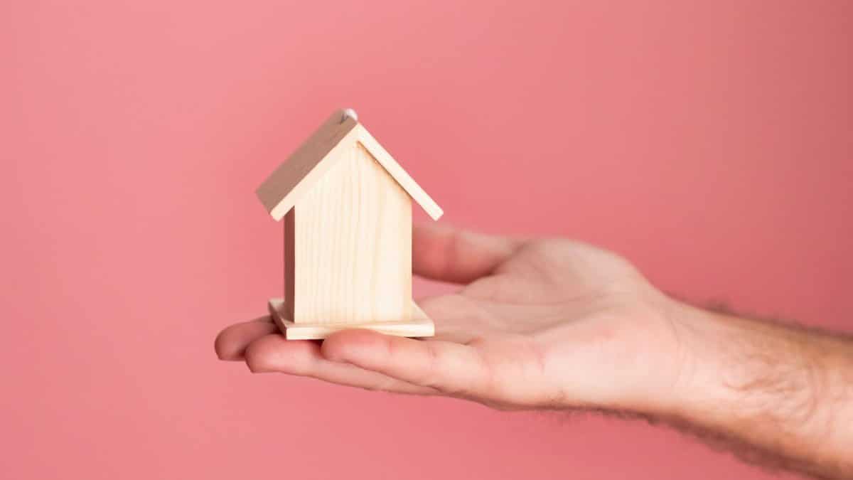 Hand holding a small wooden house model against a soft pink background, symbolizing real estate and property investment.