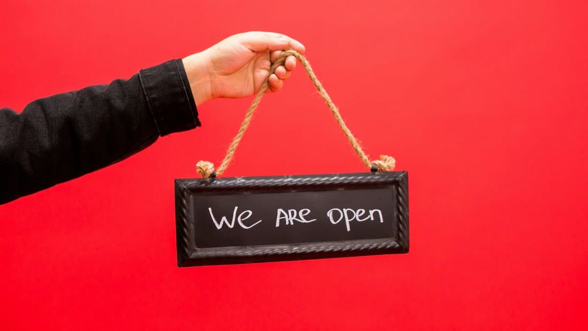 A hand holding a black sign with 'We Are Open' text, set against a vibrant red background.