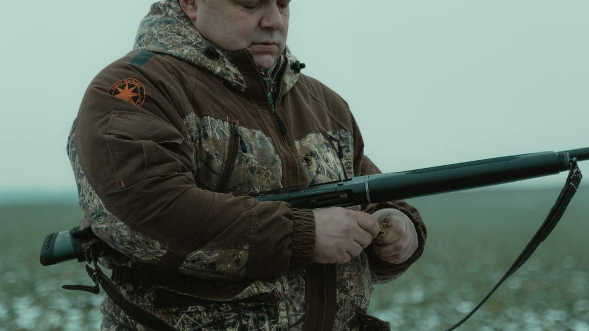 A man in camouflage reloading a rifle in an outdoor winter setting, preparing for a hunt.