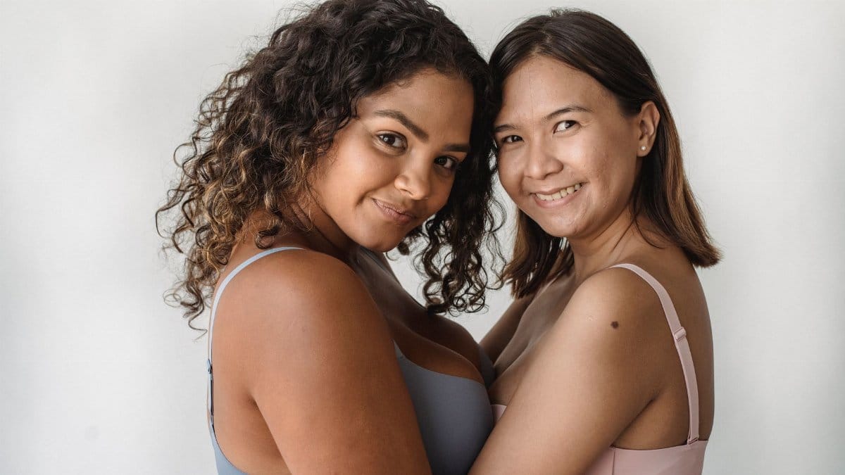Two diverse women in bras hugging and smiling against a plain background.