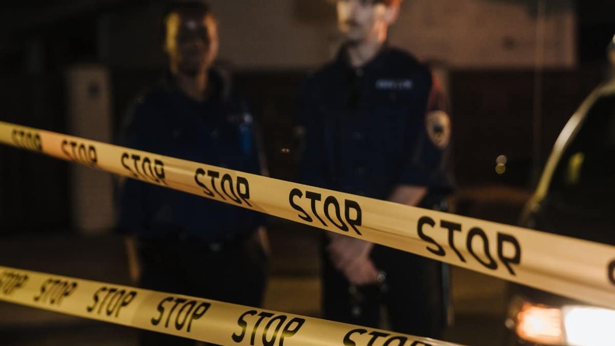 Two police officers standing behind a stop tape at night, scene partially illuminated by car headlights.