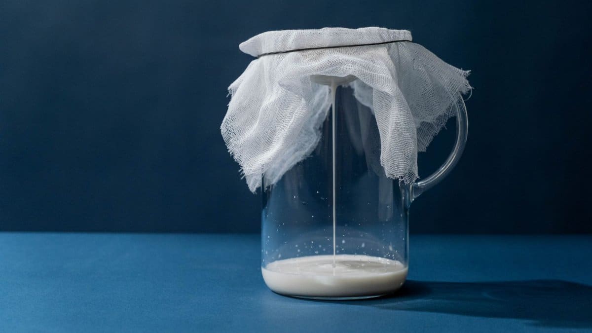 Glass pitcher with cheesecloth straining milk; minimalist still life on blue background.