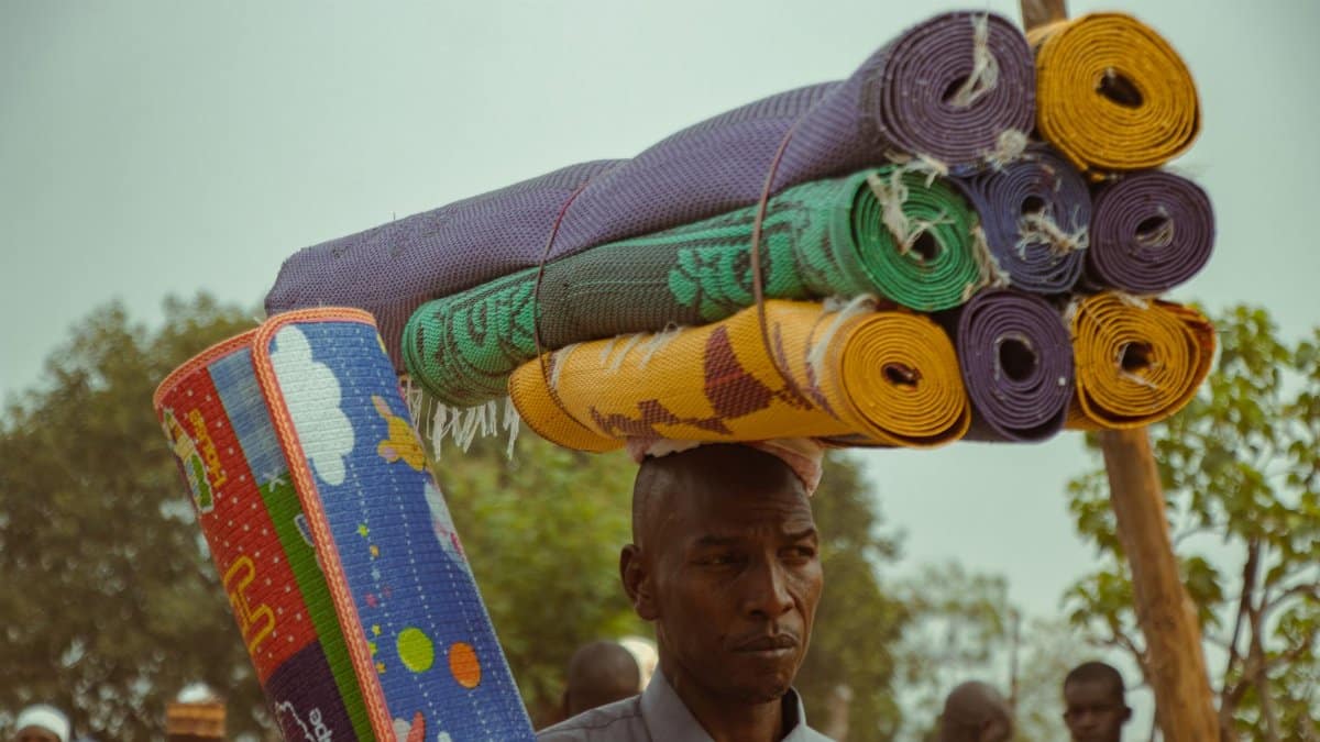 Man balancing colorful rolled mats on head at outdoor market. Summer.