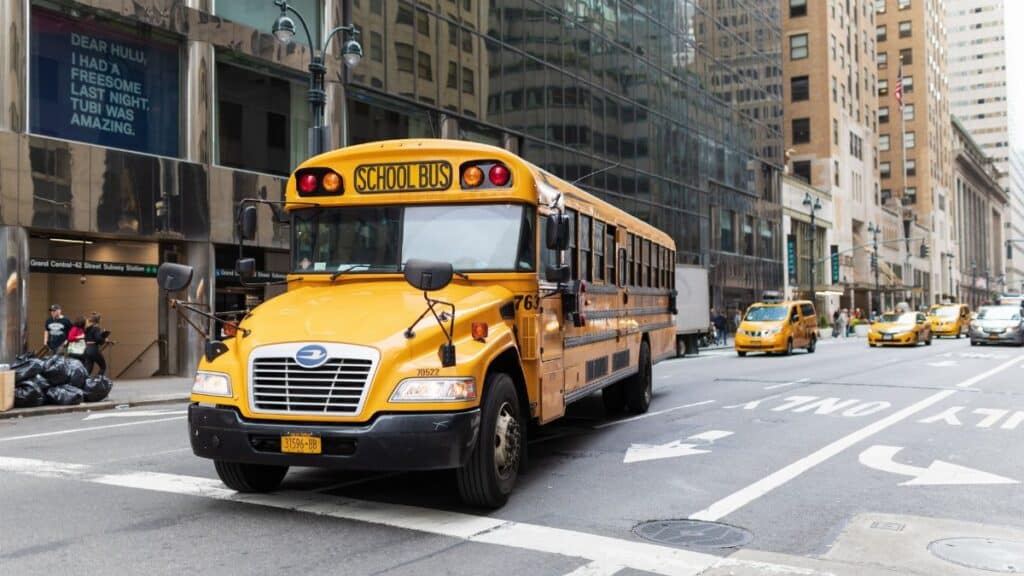 A yellow school bus navigates a busy New York City street amidst tall buildings and taxis.
